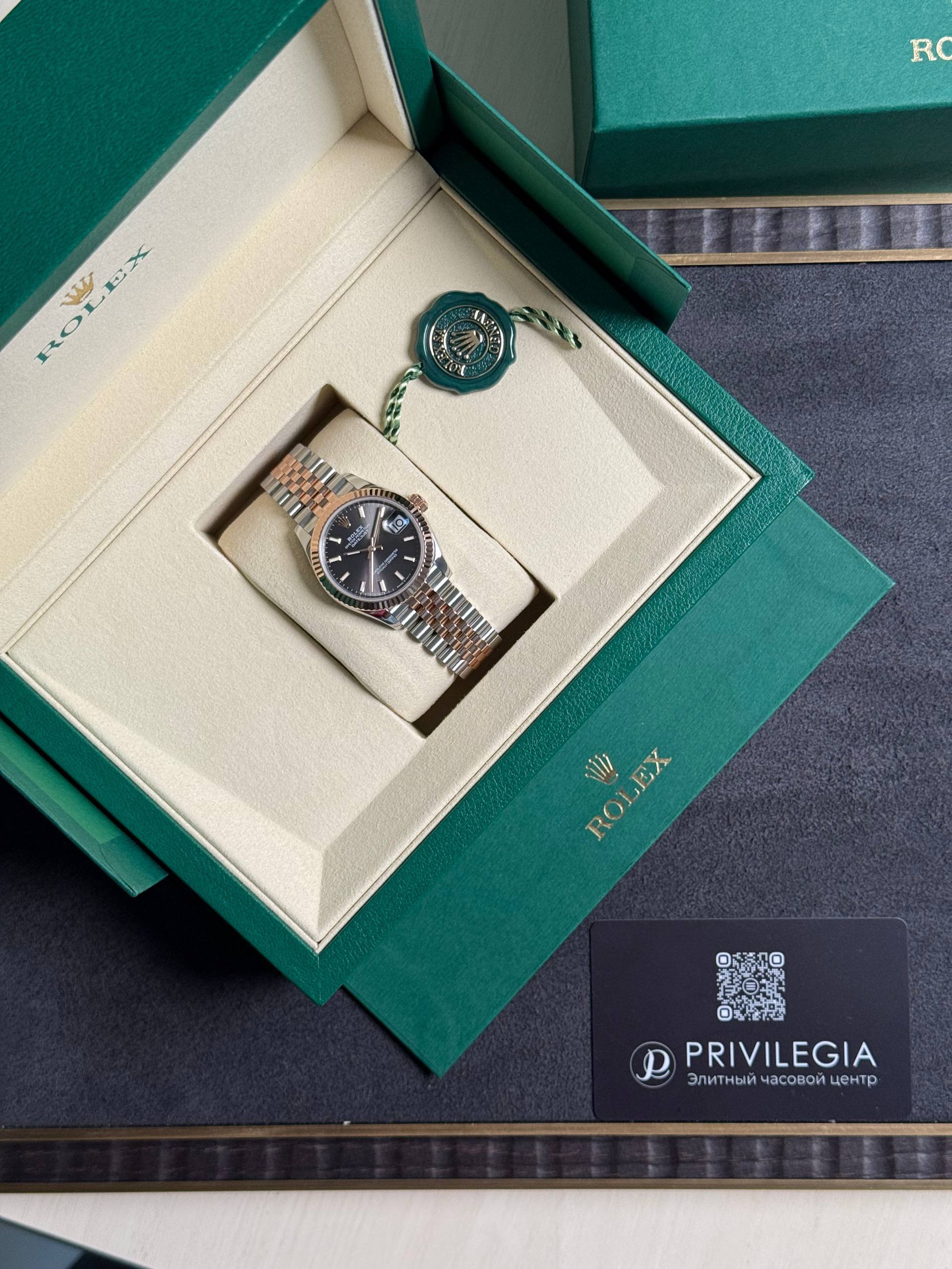 Open green Rolex box displaying a two-tone Rolex watch with black dial and date window, plus green seal tag and Privilegia card nearby.