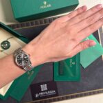 Two-tone Rolex Datejust on a wrist beside green Rolex packaging and tags, showing the timepiece clearly