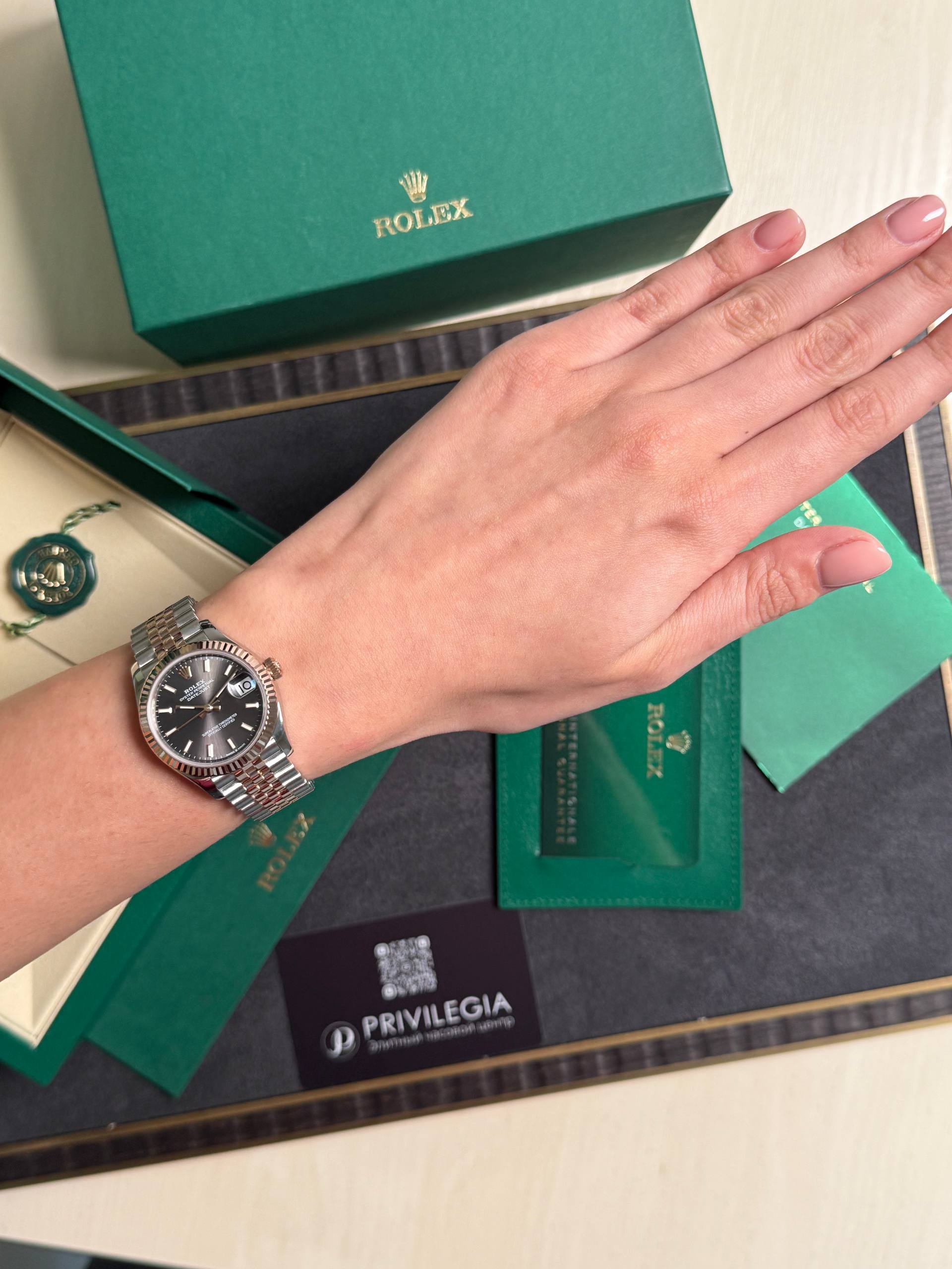 Two-tone Rolex Datejust on a wrist beside green Rolex packaging and tags, showing the timepiece clearly