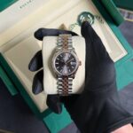 Close-up of a two-tone Rolex Datejust wristwatch in a green box, gloved hand holding it on a beige cushion