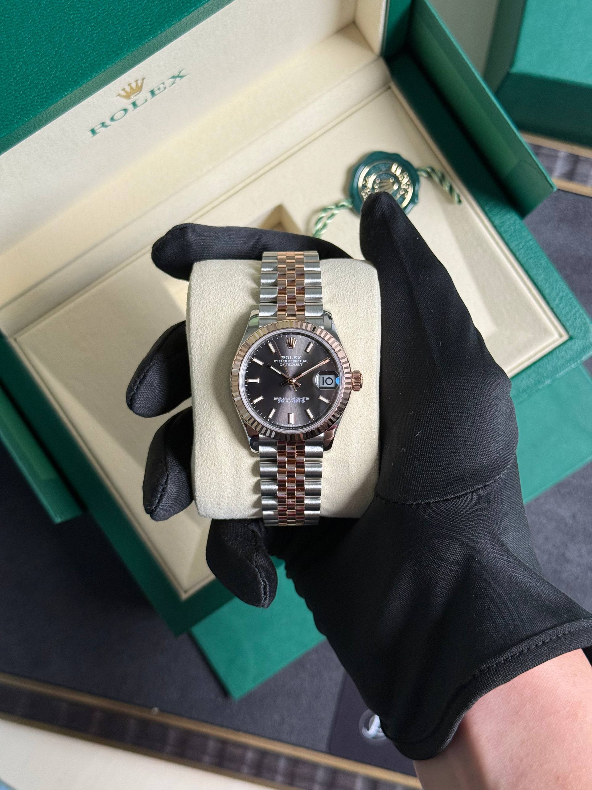 Close-up of a two-tone Rolex Datejust wristwatch in a green box, gloved hand holding it on a beige cushion