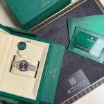 Green Rolex presentation box open to reveal a two-tone Rolex watch in a cream insert with green warranty cards nearby.