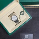Rolex wristwatch in its green presentation box with beige interior and a circular model tag, on a dark surface.