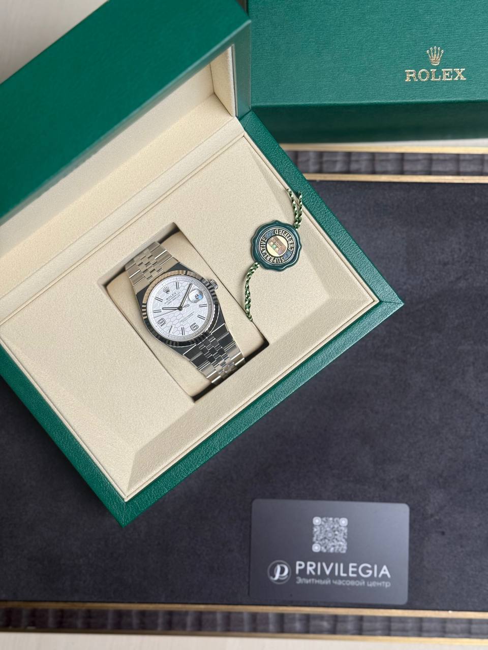 Rolex wristwatch in its green presentation box with beige interior and a circular model tag, on a dark surface.