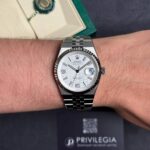 Rolex stainless steel wristwatch on a light-skinned wrist with a white patterned dial, date window at 3 o'clock, and a fluted bezel; box and card in the background.