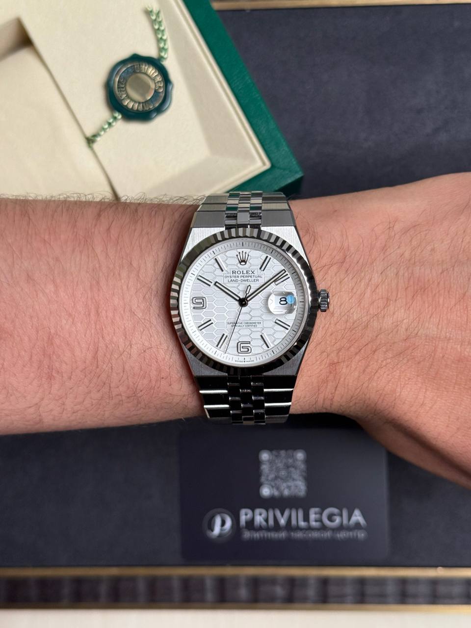 Rolex stainless steel wristwatch on a light-skinned wrist with a white patterned dial, date window at 3 o'clock, and a fluted bezel; box and card in the background.