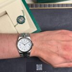 Silver Rolex watch on the left wrist with a white dial, date window, and metal bracelet, beside a green Rolex box and a seal badge.