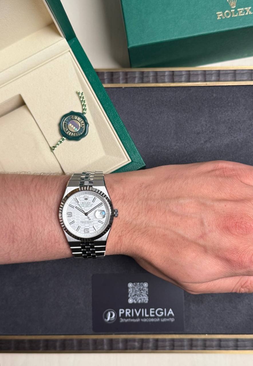 Silver Rolex watch on the left wrist with a white dial, date window, and metal bracelet, beside a green Rolex box and a seal badge.