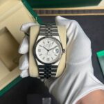 Silver Rolex watch with a fluted bezel and stainless steel bracelet, held by a white-gloved hand in front of a green Rolex box (product shot).