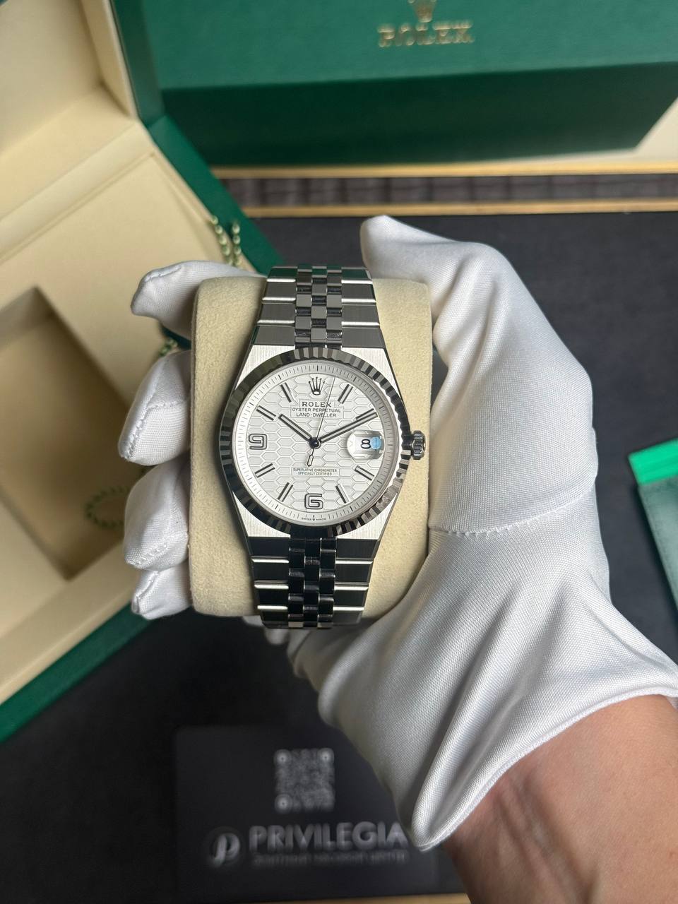 Silver Rolex watch with a fluted bezel and stainless steel bracelet, held by a white-gloved hand in front of a green Rolex box (product shot).