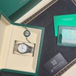Rolex watch in open green box with beige interior, alongside green card and warranty papers on a black surface