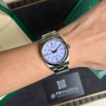 Wrist wearing a silver Rolex Oyster Perpetual with a light blue dial and metal bracelet