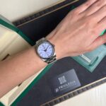 Silver metal-band wristwatch with a white dial resting on the wrist above an open green-and-black box.