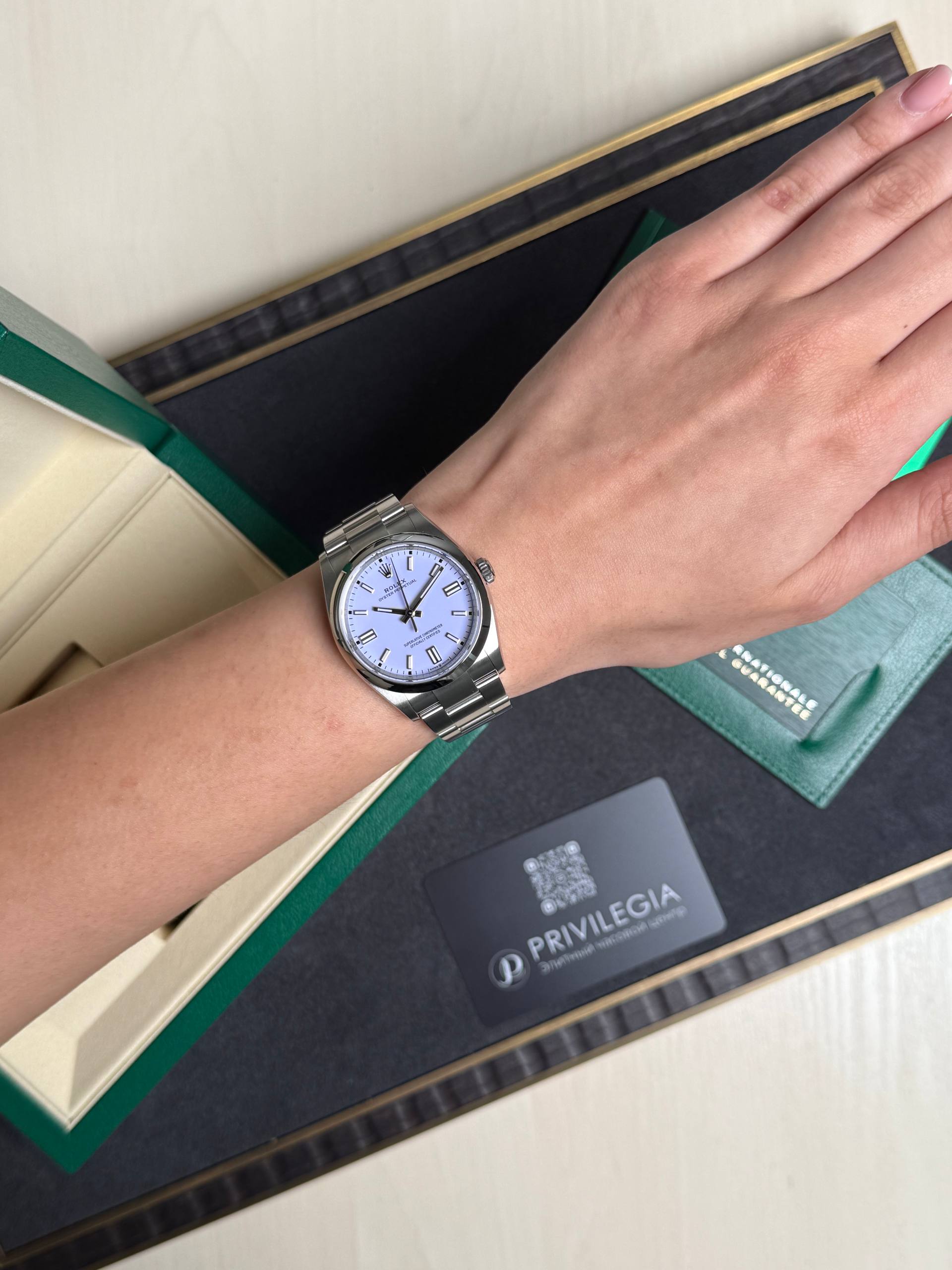 Silver metal-band wristwatch with a white dial resting on the wrist above an open green-and-black box.