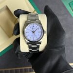 Stainless steel Rolex watch on a beige cushion, held by a gloved hand with a lavender dial showing timepieces.