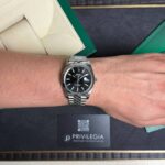 Silver Rolex watch on a man's left wrist, resting on a dark surface with green and beige packaging nearby and a Privilegia card in view.