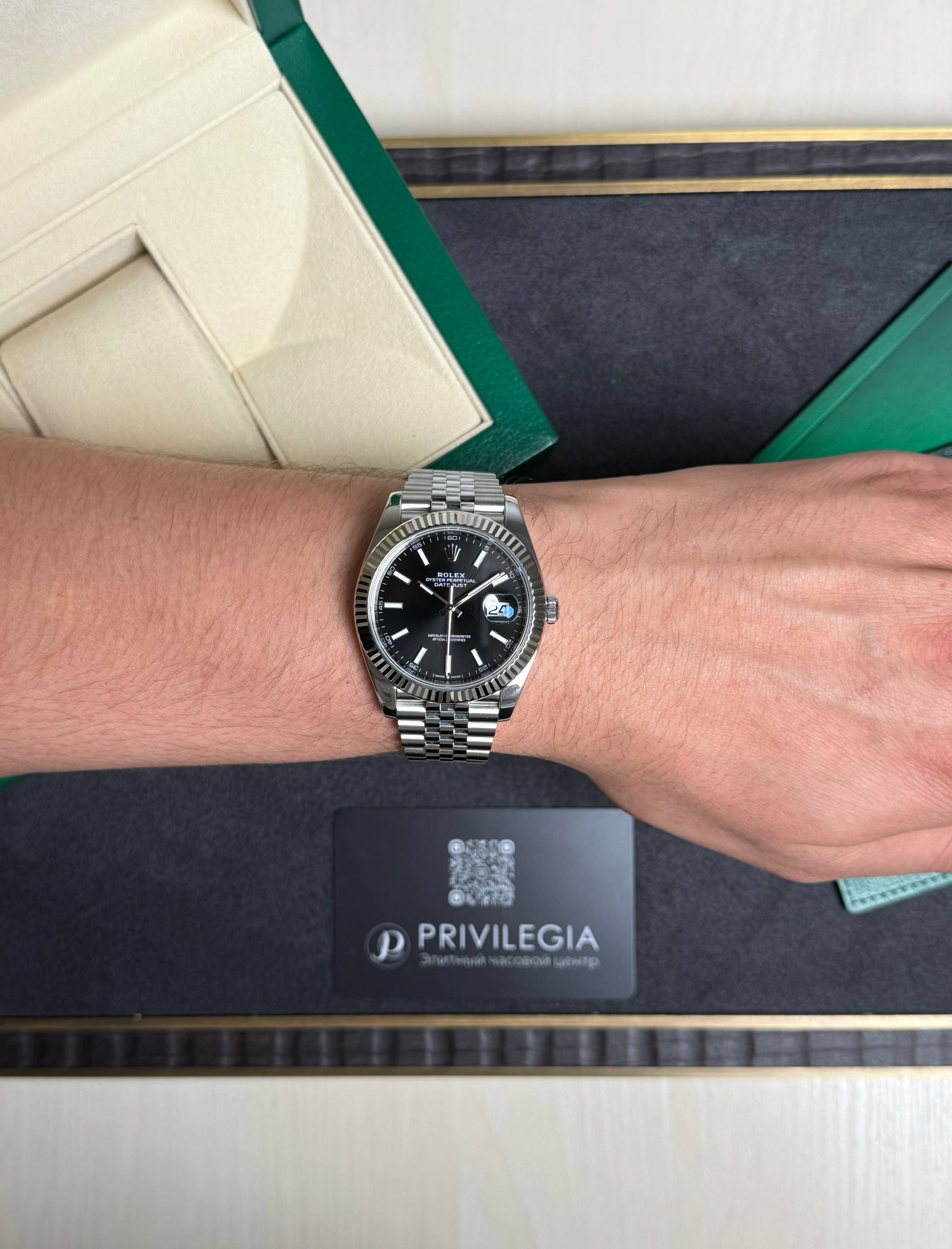 Silver Rolex watch on a man's left wrist, resting on a dark surface with green and beige packaging nearby and a Privilegia card in view.