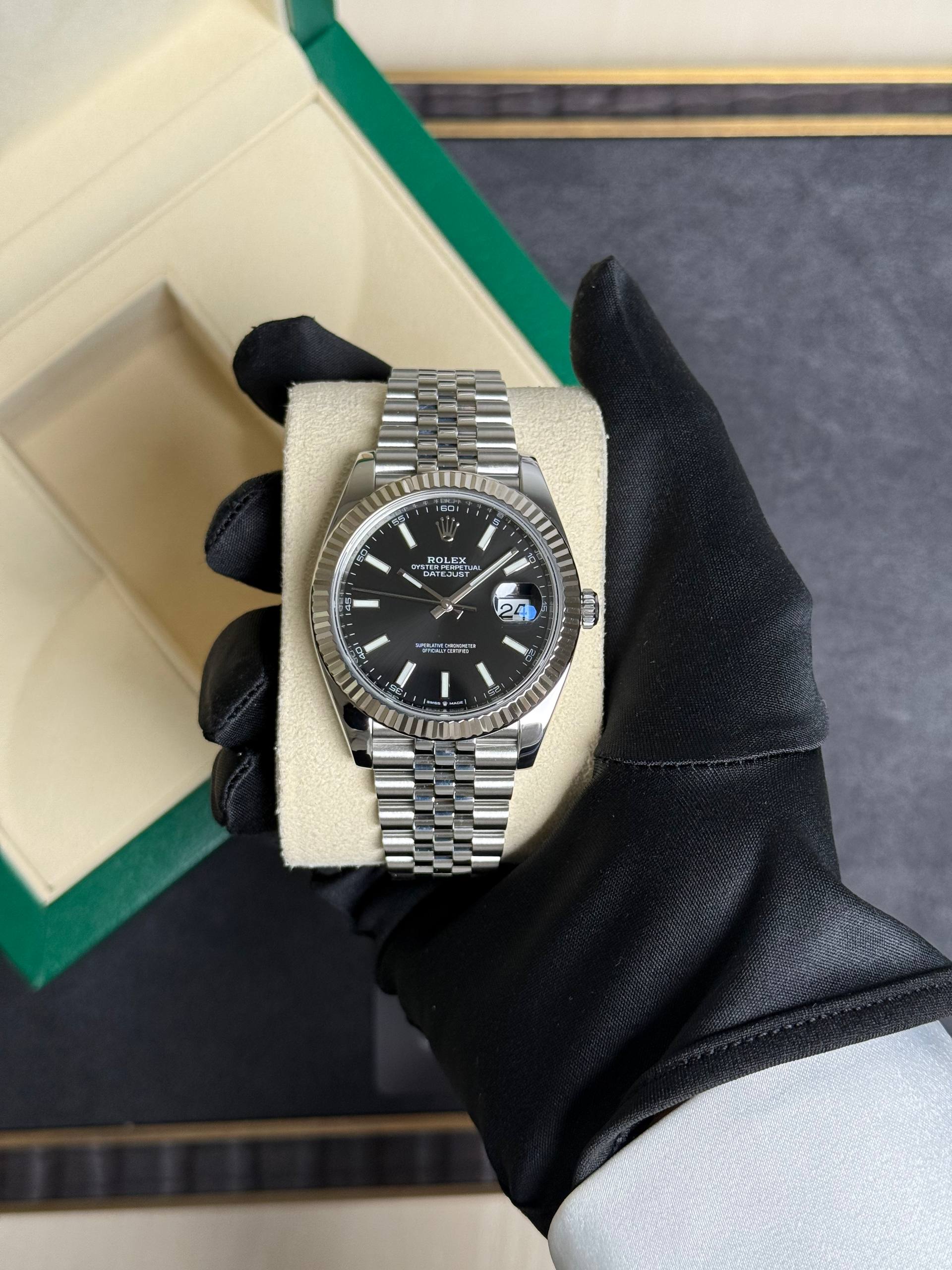 Close-up of a stainless steel Rolex Datejust watch on a beige display cushion, held by a gloved hand; black dial with silver markers and fluted bezel.