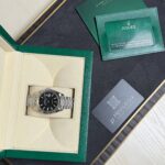 Rolex watch in a beige display box with green outer case and accompanying green warranty card and booklet pieces nearby