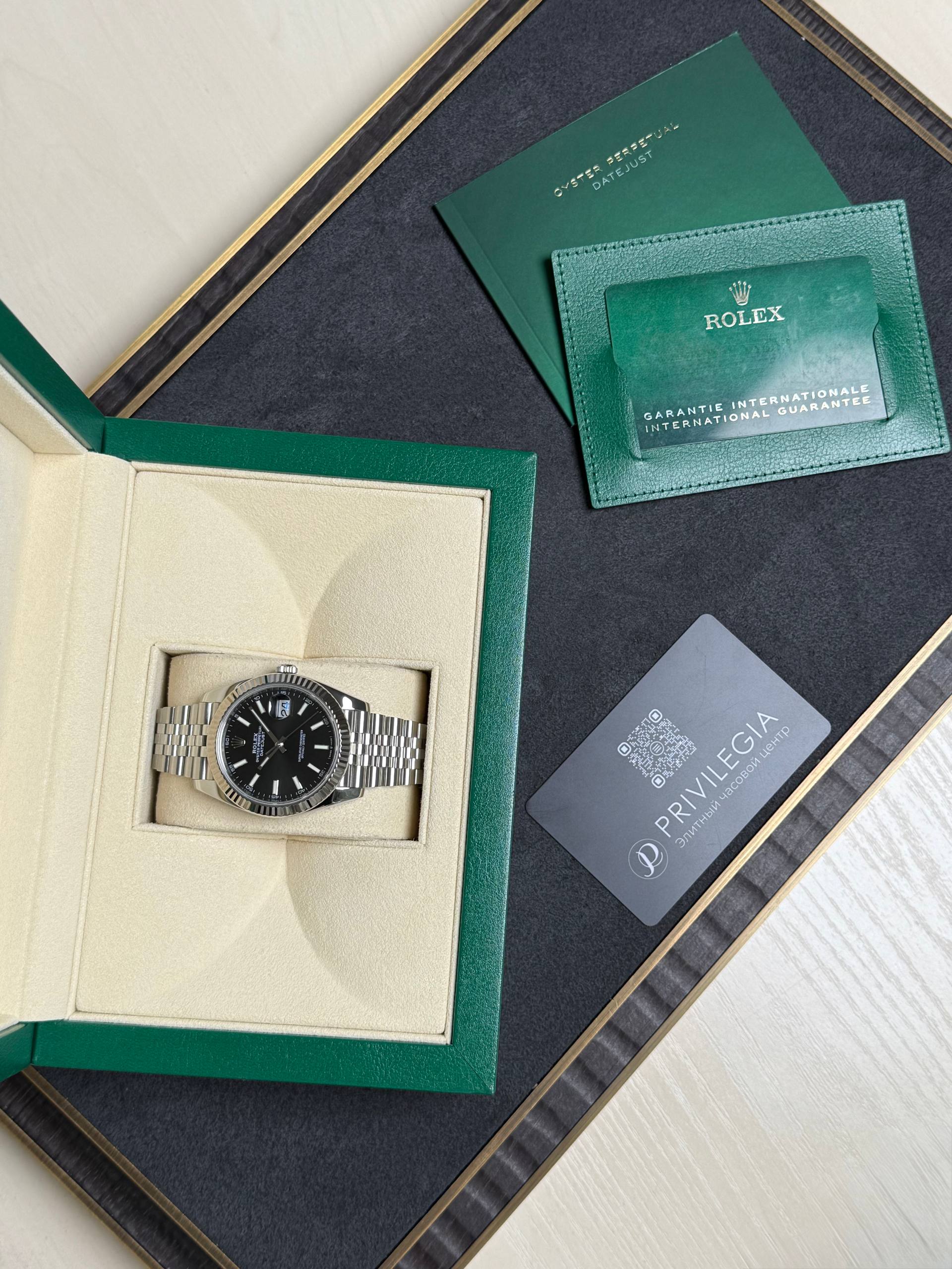 Rolex watch in a beige display box with green outer case and accompanying green warranty card and booklet pieces nearby