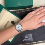 Wrist wearing a two-tone Rolex Datejust with a white dial and diamond markers, beside a green Rolex box.