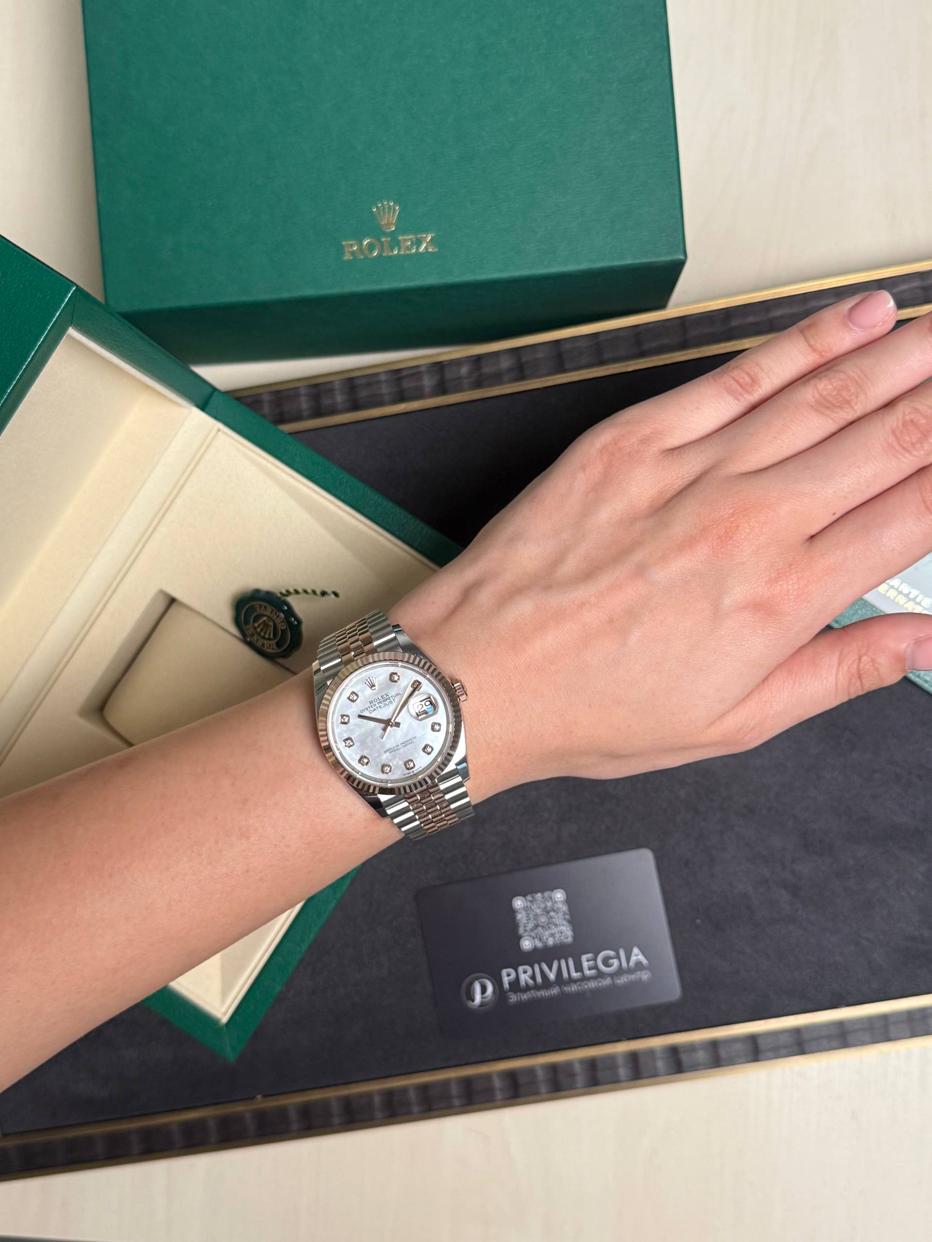 Wrist wearing a two-tone Rolex Datejust with a white dial and diamond markers, beside a green Rolex box.