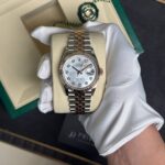 Two-tone Rolex Datejust wristwatch held in a white glove inside its beige presentation pillow, diamond hour markers and white mother-of-pearl dial.