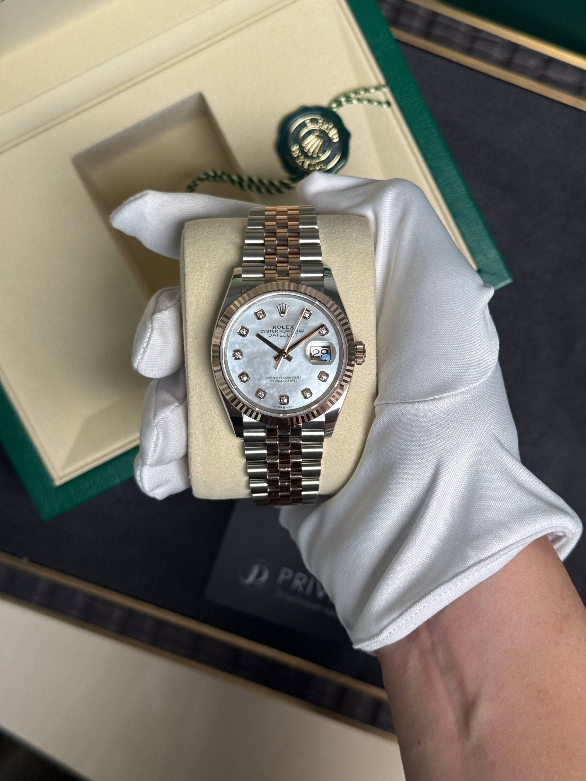 Two-tone Rolex Datejust wristwatch held in a white glove inside its beige presentation pillow, diamond hour markers and white mother-of-pearl dial.