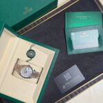 Green Rolex presentation box open to reveal a two-tone metal watch in cream interior with accessories nearby