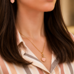 Close-up of a woman's neck showcasing a gold heart pendant necklace and matching heart earring, with a striped blouse in warm tones.