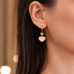 Close-up of a woman's ear wearing a gold double-heart dangle earring: a small gray heart above a larger pink heart.