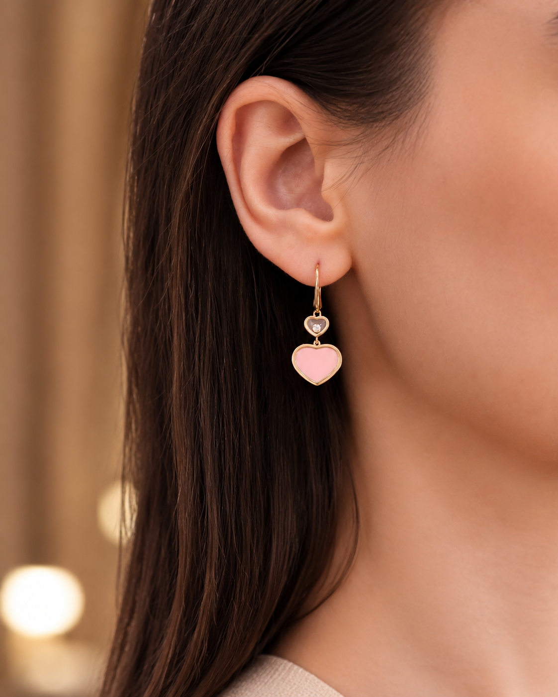 Close-up of a woman's ear wearing a gold double-heart dangle earring: a small gray heart above a larger pink heart.