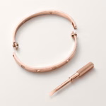 Open rose-gold bangle bracelet with crystal accents, accompanied by a small precision screwdriver on a pale background.