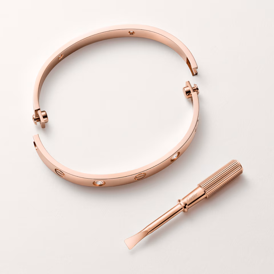 Open rose-gold bangle bracelet with crystal accents, accompanied by a small precision screwdriver on a pale background.