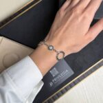 Woman's wrist wearing a silver bracelet with circular linked charms, including a lucent stone centerpiece; background shows a dark jewelry box and branding 'BVLGARI'.