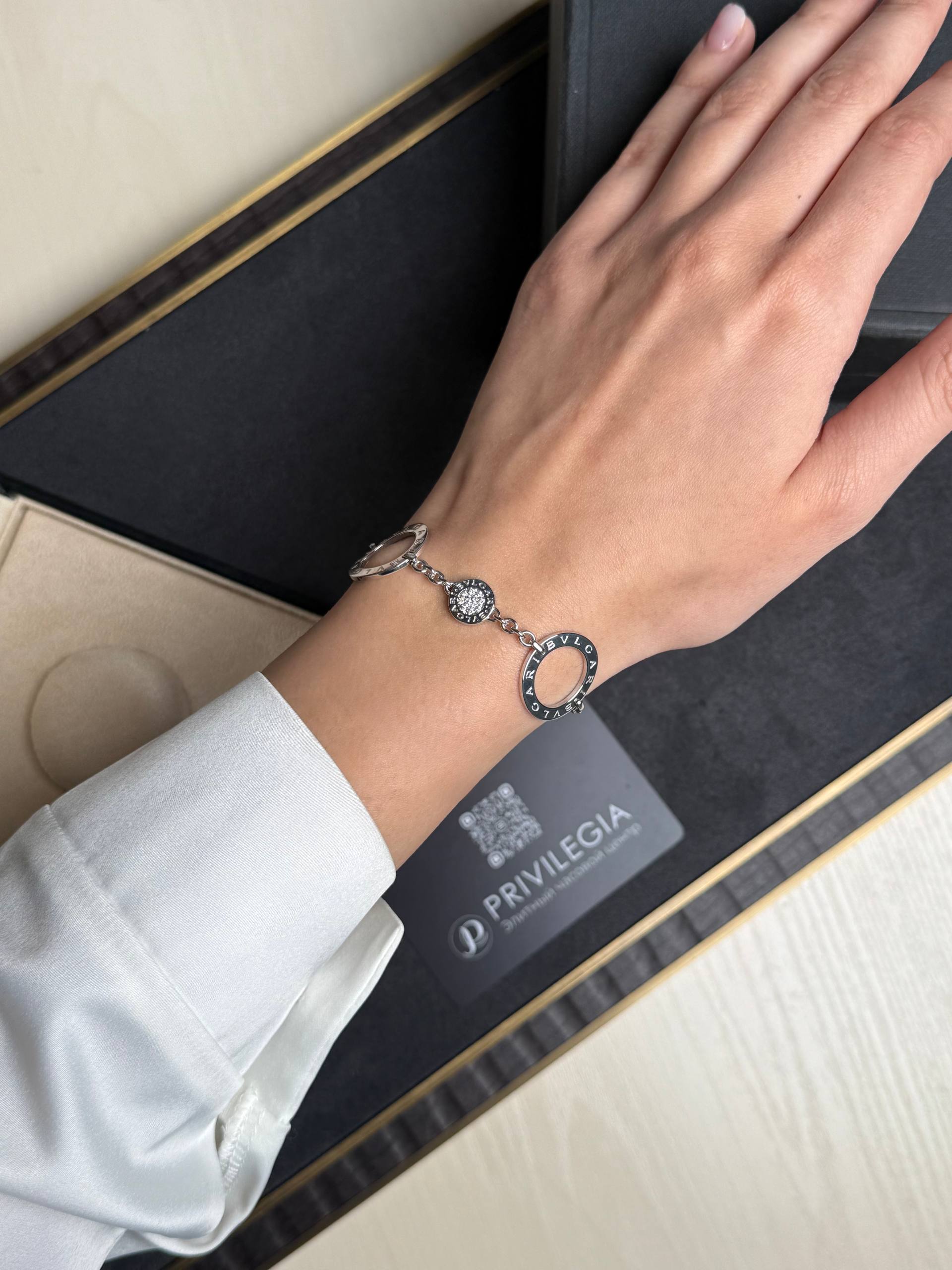 Woman's wrist wearing a silver bracelet with circular linked charms, including a lucent stone centerpiece; background shows a dark jewelry box and branding 'BVLGARI'.
