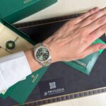 Wrist wearing a two-tone Rolex watch with a green dial, resting on green Rolex packaging boxes and a Privilegia card nearby.