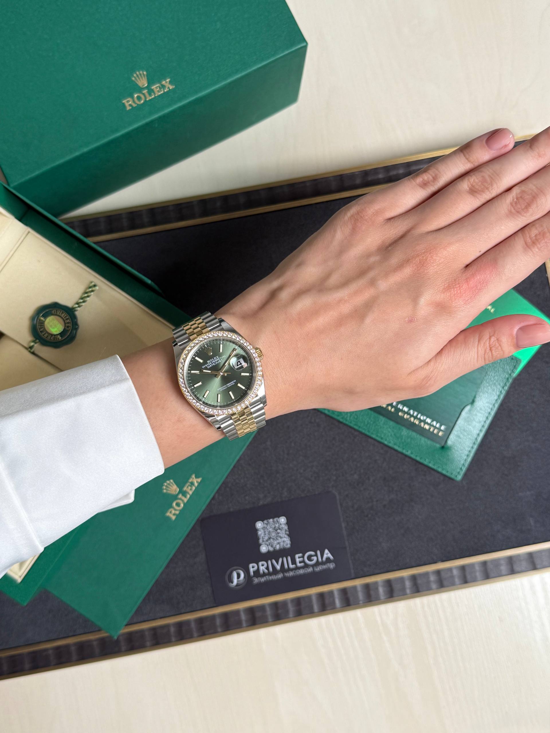 Wrist wearing a two-tone Rolex watch with a green dial, resting on green Rolex packaging boxes and a Privilegia card nearby.