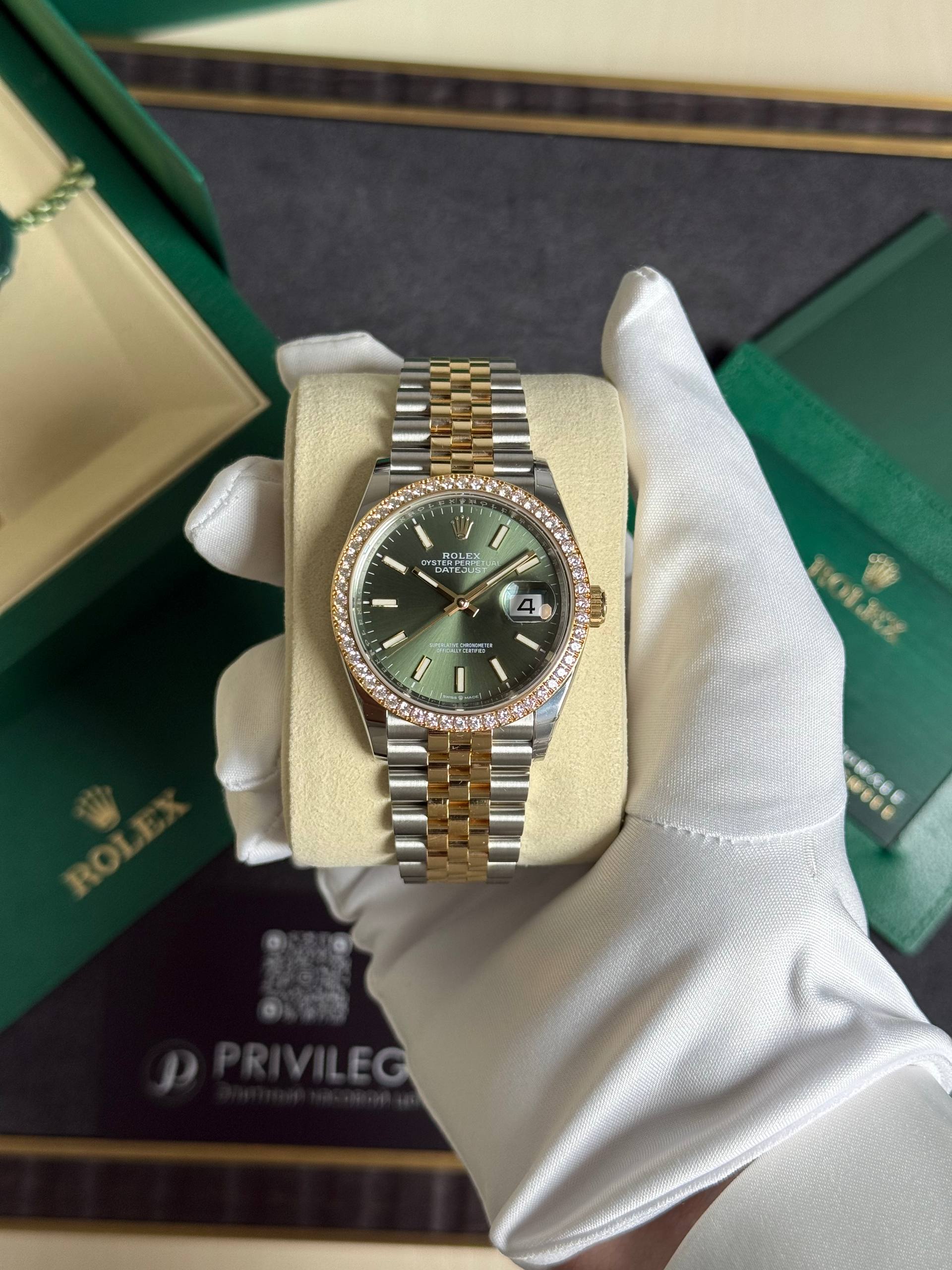 Two-tone Rolex Datejust with green dial and diamond bezel held in white gloves on a display cushion