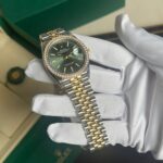 Rolex Datejust watch with emerald-green dial, diamond bezel, two-tone gold and stainless steel bracelet, held by white gloves.