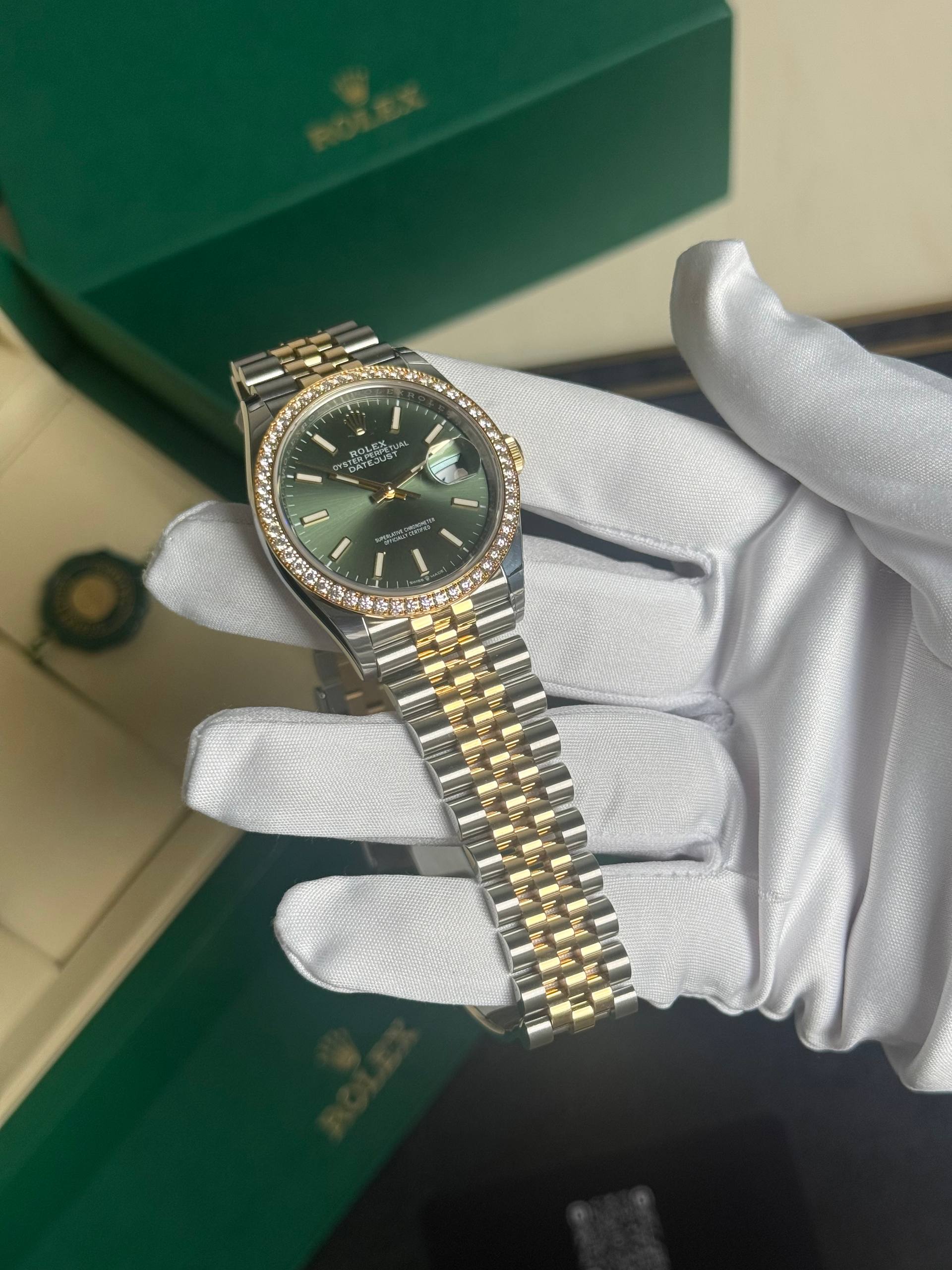 Rolex Datejust watch with emerald-green dial, diamond bezel, two-tone gold and stainless steel bracelet, held by white gloves.