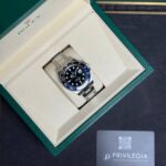 Rolex watch in an open green presentation box with beige interior, showing a stainless steel bracelet and dark dial with date window; Privilegia card nearby.