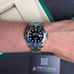 Wrist with a stainless Rolex GMT‑Master II watch, black dial and blue/red bezel, silver bracelet, packaging in the background.