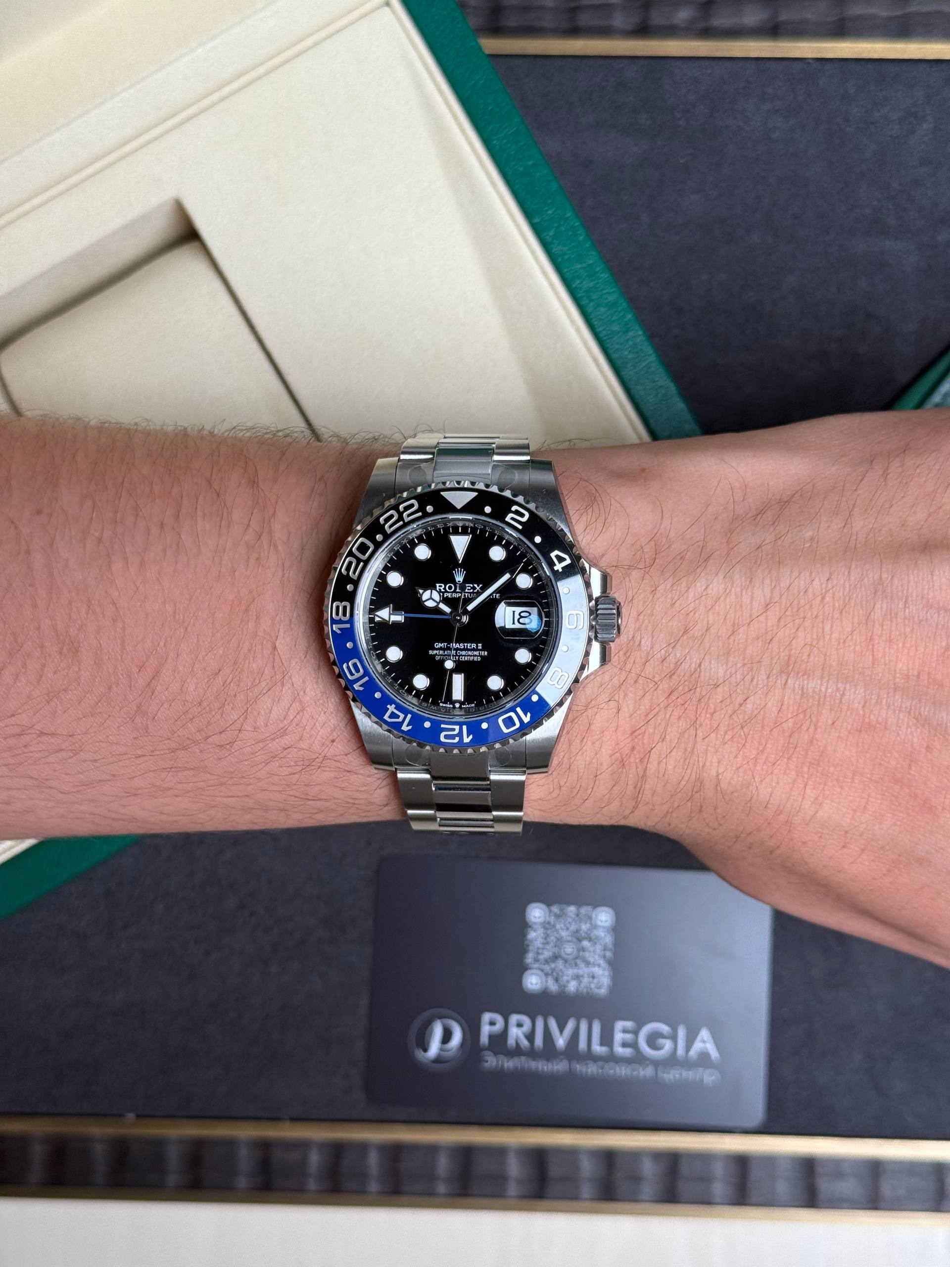 Wrist with a stainless Rolex GMT‑Master II watch, black dial and blue/red bezel, silver bracelet, packaging in the background.