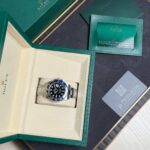 Rolex watch in an open beige interior box with green outer case and accompanying warranty cards and papers nearby.