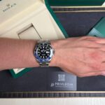 Wrist wearing a stainless steel Rolex GMT-Master II with blue/black bezel, open green Rolex box in the background.