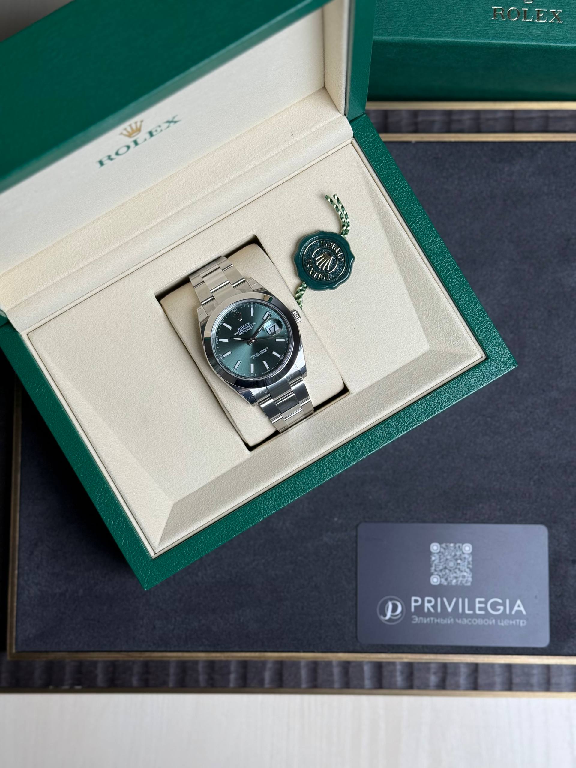 Rolex watch in an open green box with a cream interior, green seal tag nearby.