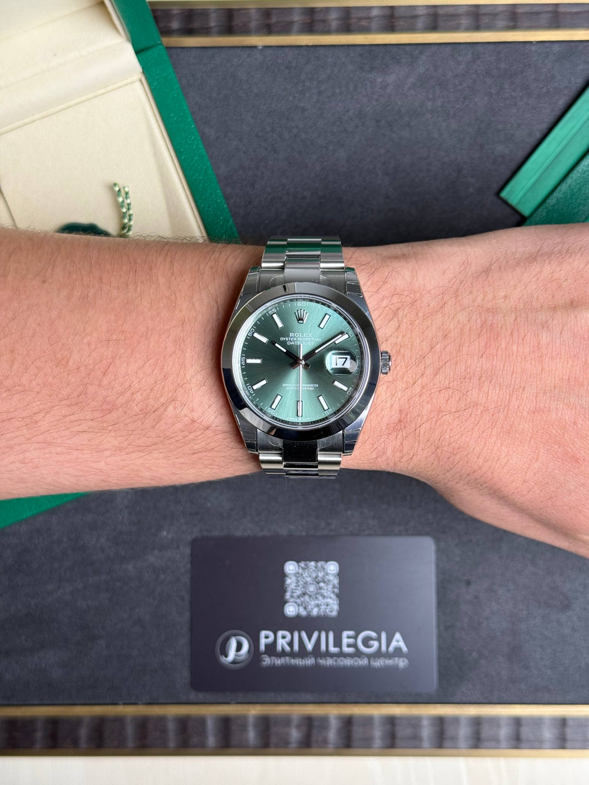 Wrist wearing a stainless steel Rolex with green dial and date window on the bracelet roll hubbing a dark surface.