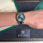 Silver Rolex watch with a green dial on a metal bracelet worn on the wrist, with green packaging in the background.