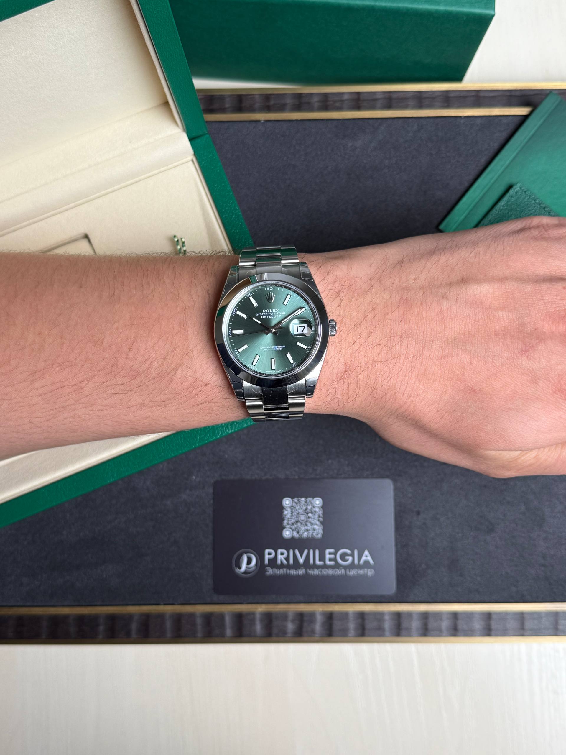Silver Rolex watch with a green dial on a metal bracelet worn on the wrist, with green packaging in the background.